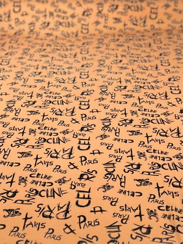 Close-up of peach-colored fabric covered with a repeating pattern of black fashion brand names such as Paris, Celine, and Chanel, with logos.
