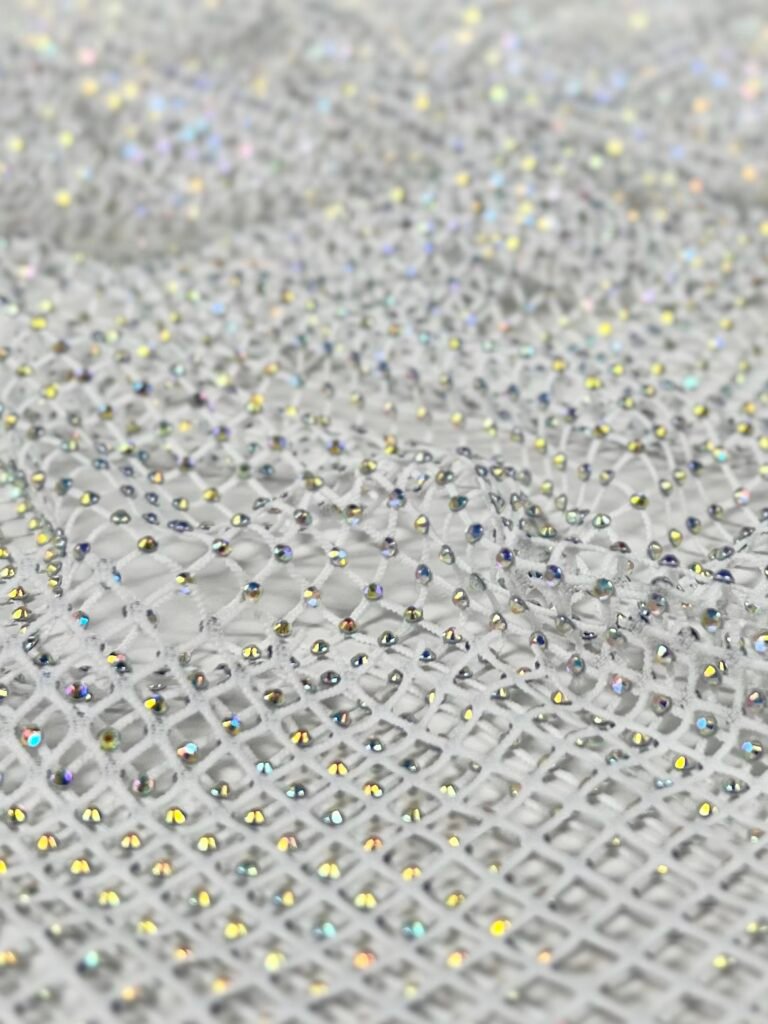 Close-up of white mesh fabric densely studded with tiny iridescent rhinestones, reflecting multicolored light.