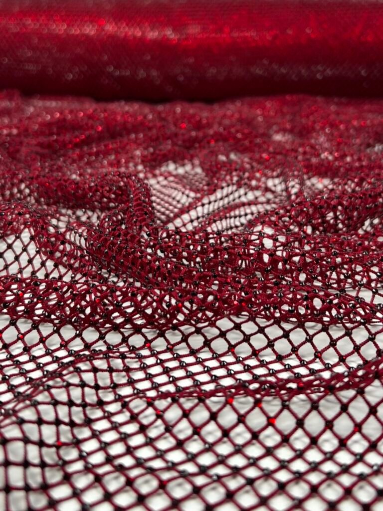 Close-up of deep red lace fabric with an open mesh pattern and tiny beads along the threads on a light background, showing intricate texture up close
